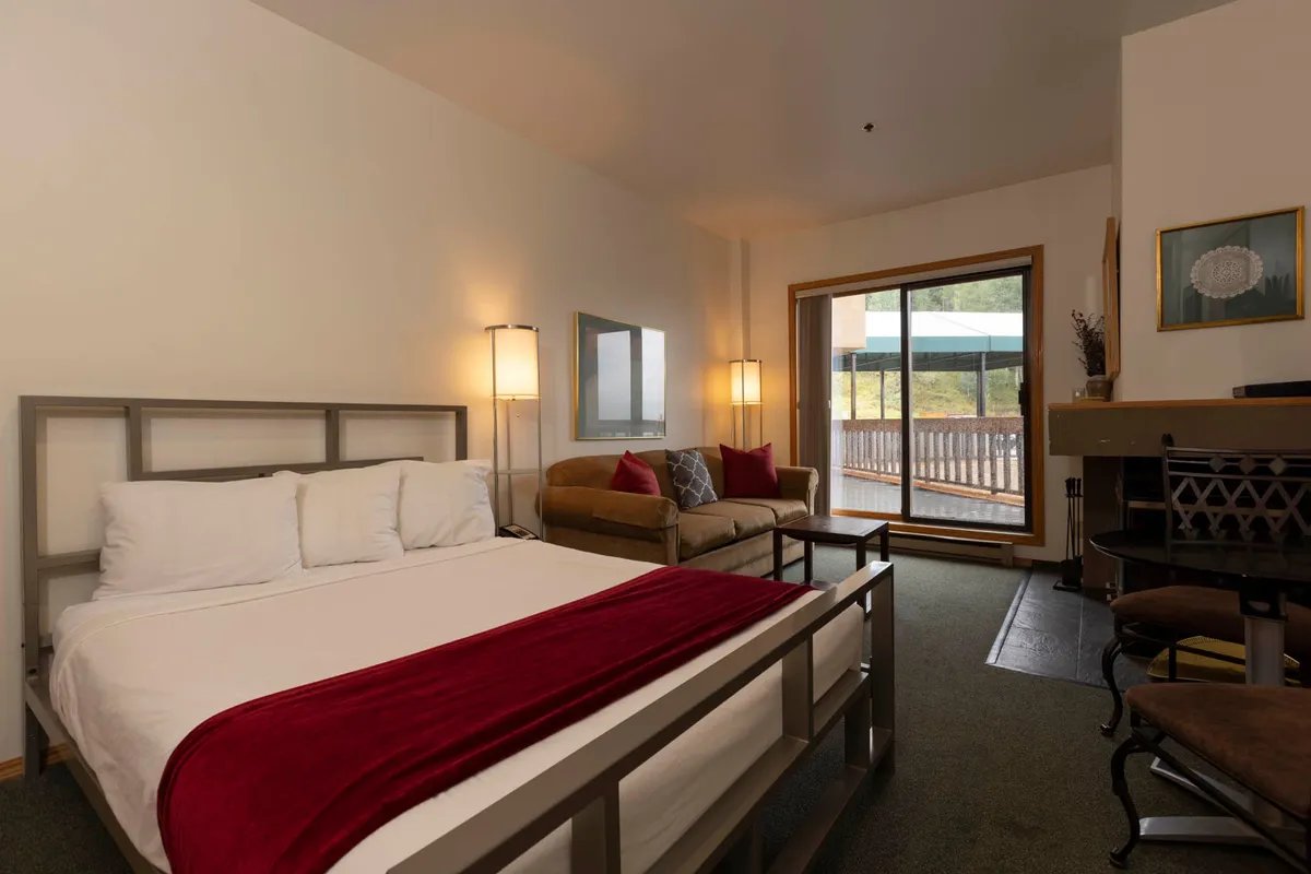 The image showcases a cozy condo interior with a plush bed, a comfortable sofa positioned near a large window, and a wood-burning fireplace as the focal point. Warm sunlight filters in, inviting guests to relax, read, or simply enjoy the scenic mountain views beyond.