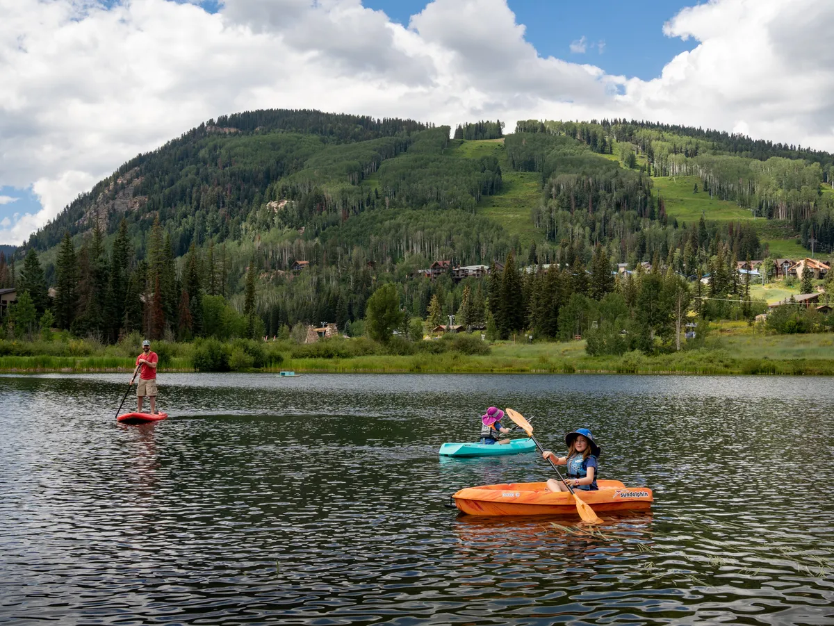 This image captures a peaceful lake scene with a paddleboarder and several kayakers moving across calm water, all set against a breathtaking mountain backdrop. The reflections on the water add to the sense of serenity and natural beauty, suggesting a tranquil environment ideal for unwinding and connecting with nature.