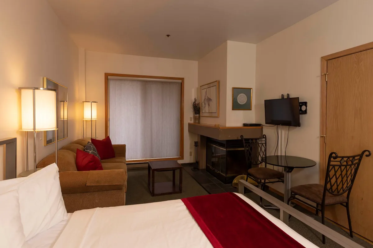 A warm, inviting living space featuring a plush bed with a red accent blanket, a comfortable sofa adorned with cushions, a flickering fireplace, and a compact dining table set for shared meals. The setting suggests a cozy retreat after a day of mountain adventures, with multiple zones for relaxation and family togetherness.