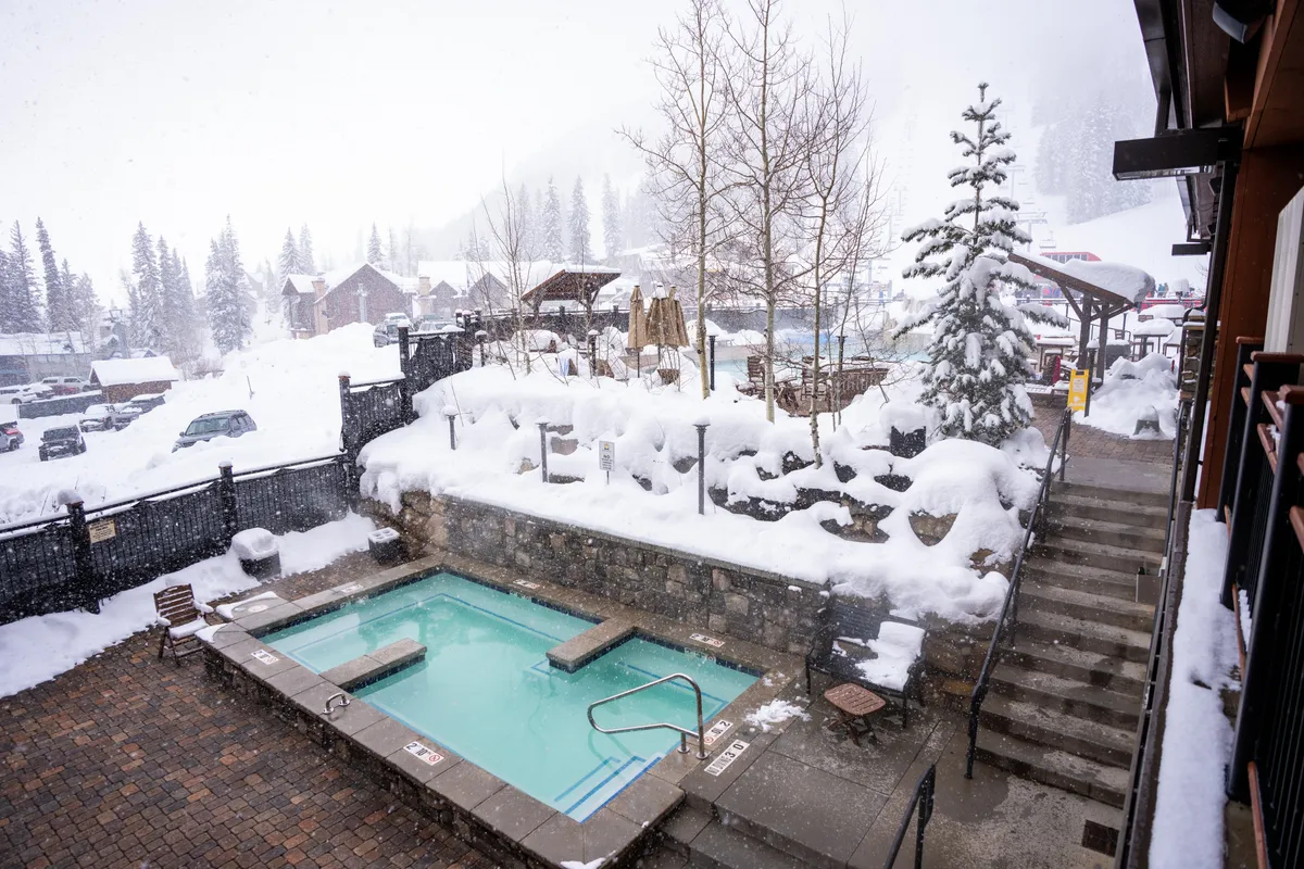 This image captures a shared outdoor hot tub set against a snowy landscape, with surrounding trees adding to a tranquil, resort-like atmosphere. The outdoor seating area and the visible contrast between the steamy hot tub and the crisp, white snow create a sense of luxurious relaxation.