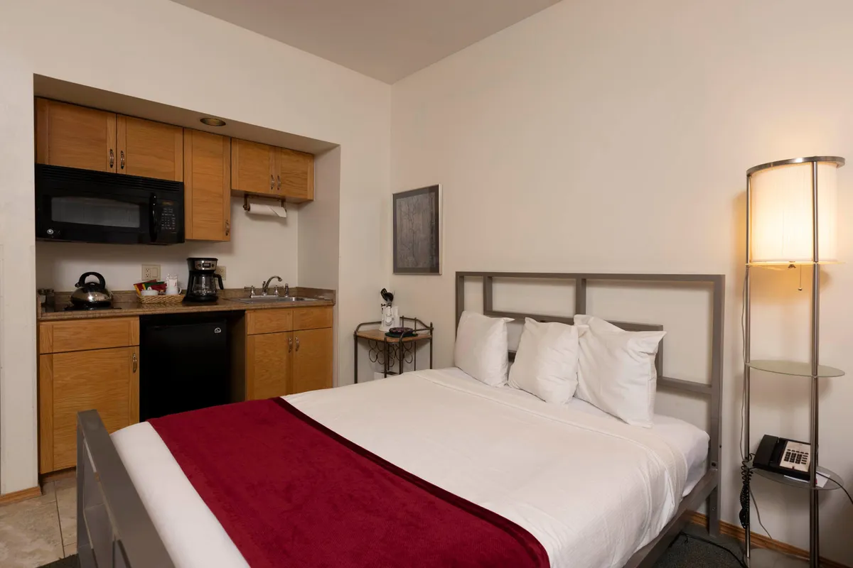 This image features a cozy interior view with a bed adorned in white linens and a red accent, a compact kitchenette area equipped with essential appliances, and a softly lit lamp. The kitchenette is clearly visible beside the sleeping area, emphasizing convenience and the ability to prepare meals or snacks close at hand.