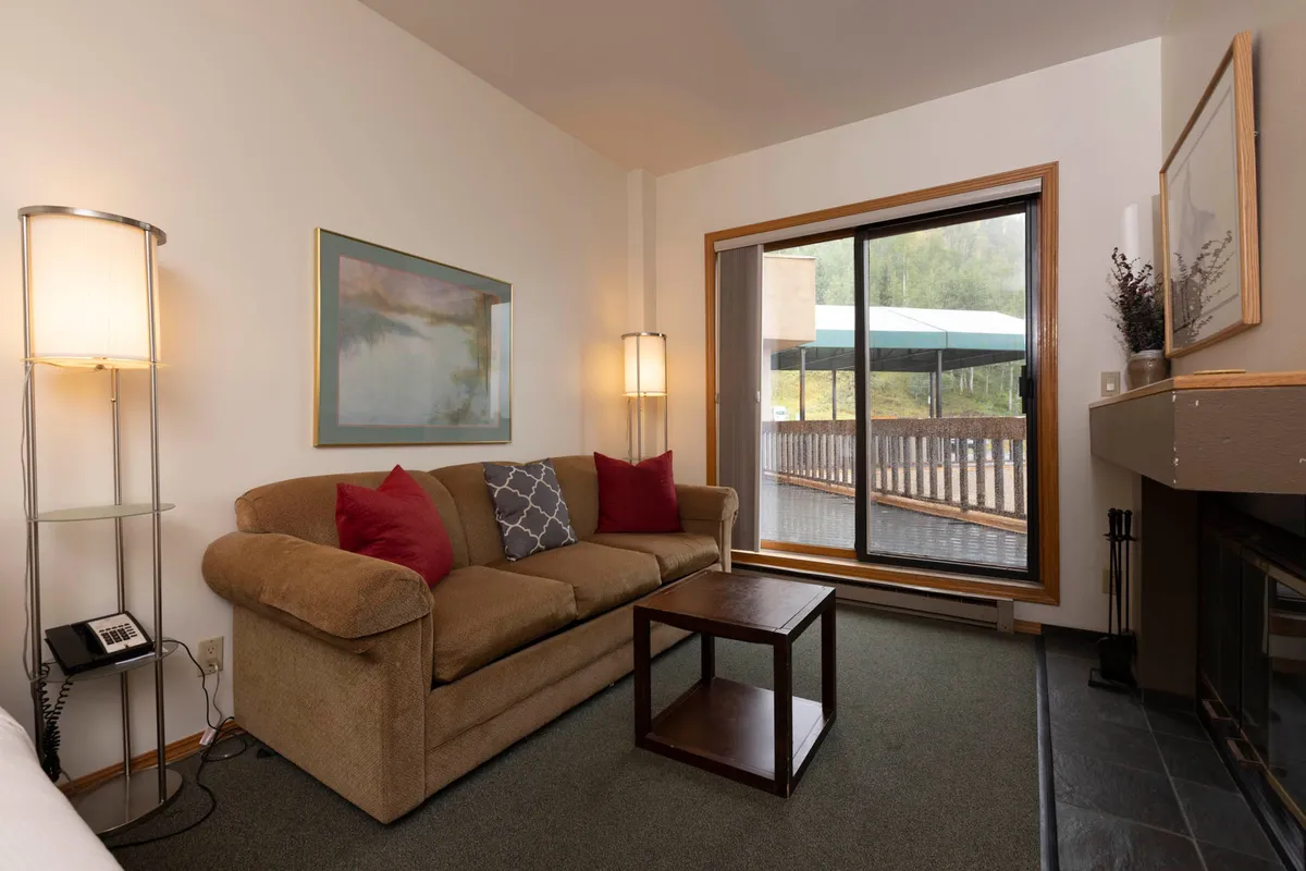 A cozy living area featuring a plush sofa and coffee table, with a large window allowing in natural light. The focal point is a beautiful wood-burning fireplace, with tasteful artwork and decor adding warmth and character. The room invites relaxation, reading, and socializing, embodying a welcoming, mountain-inspired ambiance.
