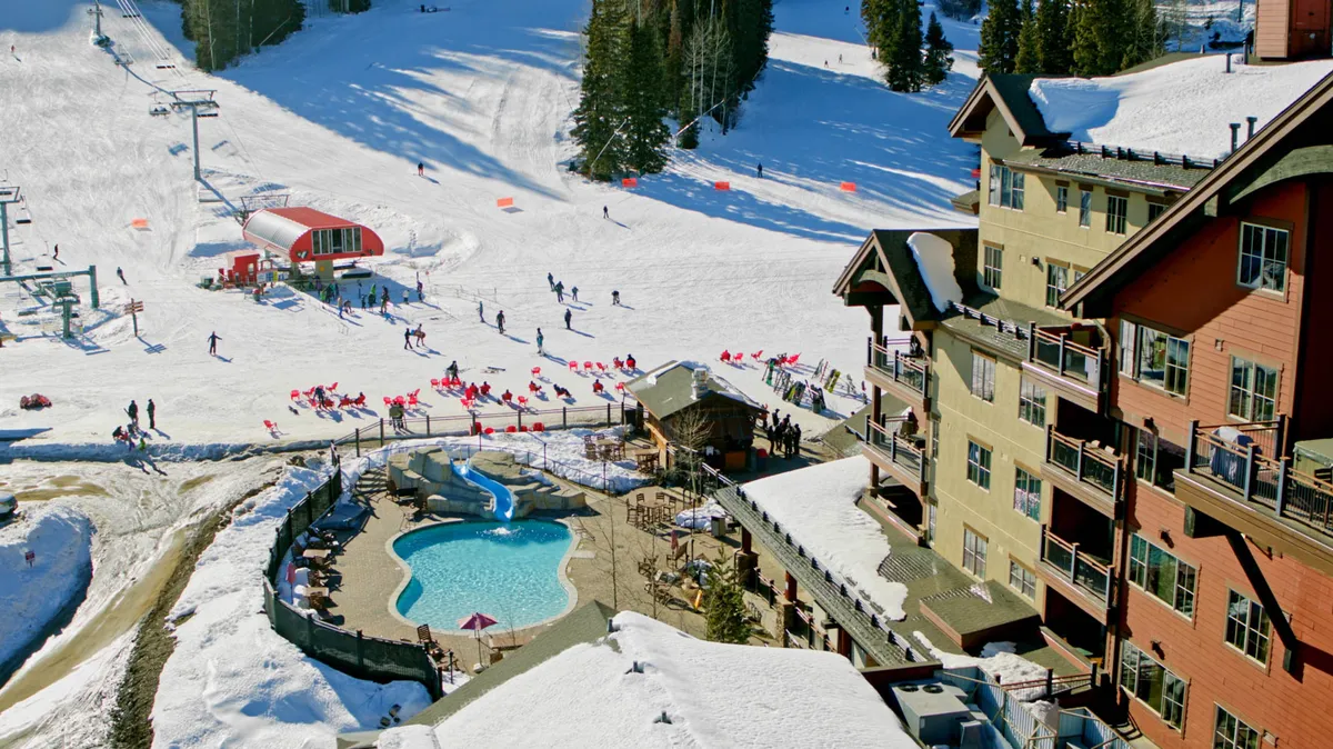 An exterior image captures both the lively ski slopes and the inviting pool area adjacent to the lodge. Skiers are visible enjoying the runs, while others gather or relax near the pool, all set against a stunning sunset over snow-dusted mountain peaks. The lodge exterior highlights the social, amenity-rich experience central to the property.