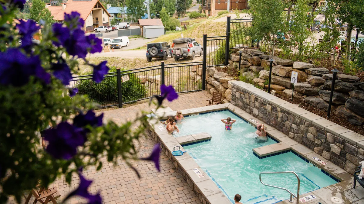 An inviting outdoor heated pool bordered by lush flowering plants, with people enjoying the water under bright sunlight. The cheerful vibe and social activity highlight the pool as a lively hub for relaxation and connection.