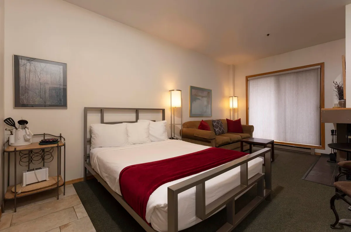 This image features a bed draped with a red blanket, a comfortable sofa, and tasteful artwork on the wall. Natural light streams softly through the blinds, enhancing the tranquil, inviting mood. The arrangement suggests a space for quiet moments, such as reading or enjoying a morning coffee.