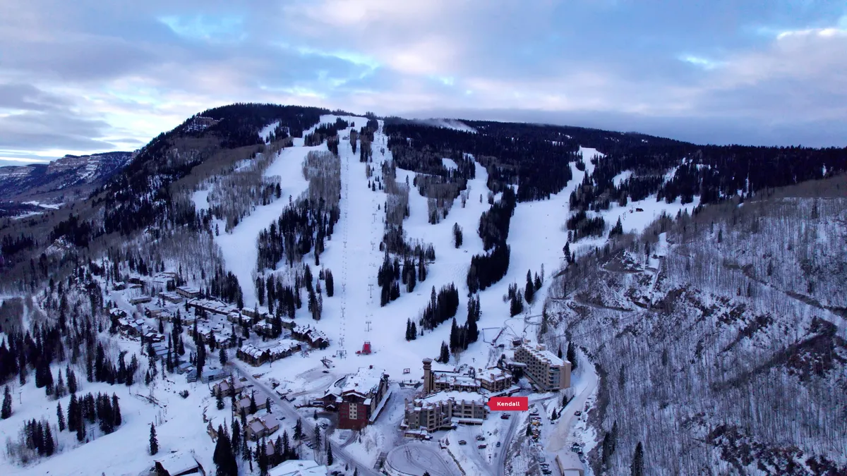A panoramic view of the village resort setting with snow-covered ski slopes, tall fir trees dusted with snow, and clusters of lodge-style resort buildings nestled in the mountains. The scene is serene, yet hints at adventure, with traces of ski runs and snowboarding activity in the landscape. The golden hues of sunset bathe the area, creating an inviting atmosphere for outdoor exploration.