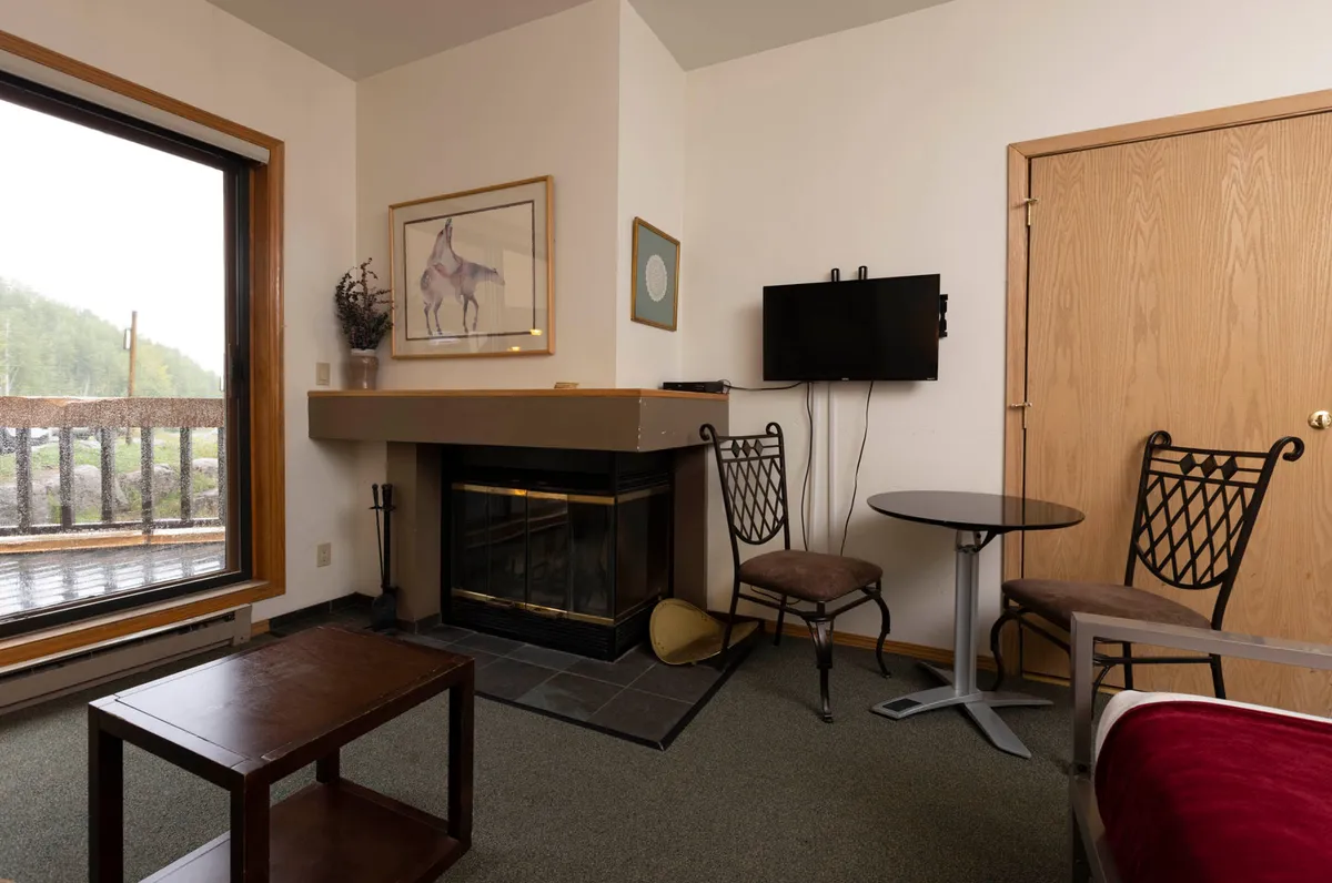 A thoughtfully arranged living space with a prominent fireplace, comfortable furniture, and large windows offering a glimpse outside. Artwork and soft lighting contribute to a warm, intimate atmosphere ideal for relaxing by the fire with a book or enjoying quiet evenings together.